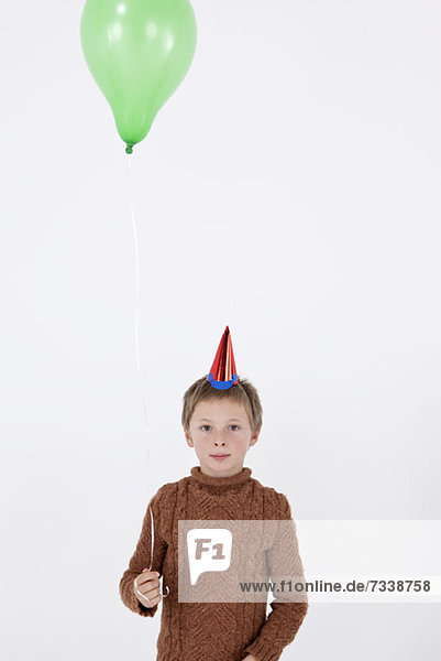 Ein ruhiger Junge  der einen Partyhut trägt und einen Ballon hält.