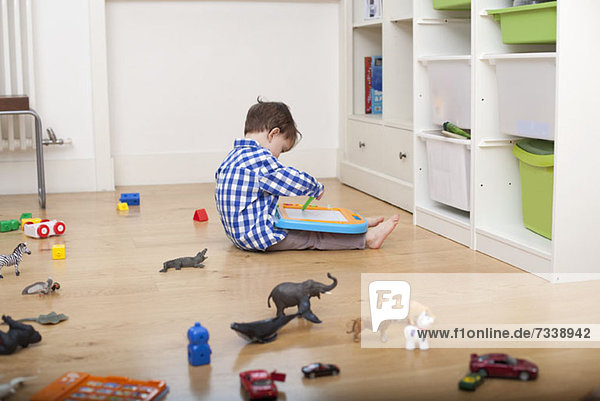 Ein Junge sitzt auf dem Boden und zeichnet auf einer Tafel  umgeben von verschiedenen Spielsachen.