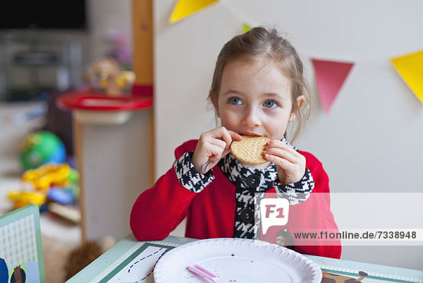 Ein kleines Mädchen  das an einem Tisch sitzt und einen Bissen aus einem Keks nimmt.