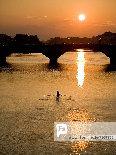 Man rowing on a body of water with a bridge and setting sun in the background.