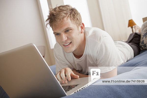 Smiling man lying on bed using laptop