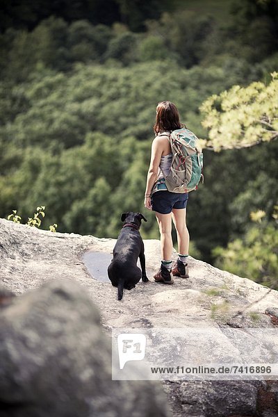 A female hiker and her dog look out over dense forest from a clifftop viewpoint.