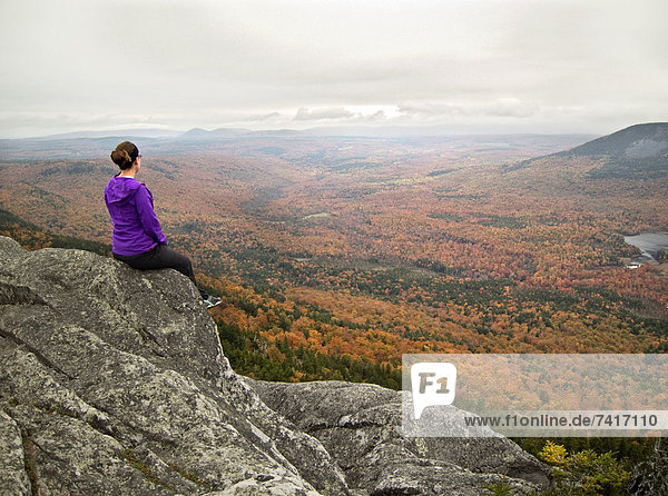 A young female hiker views vibrant fall foliage from the rocky summit of a New England peak.