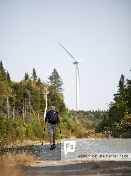 A man hikes along a trail with a wind turbine looming in the background.