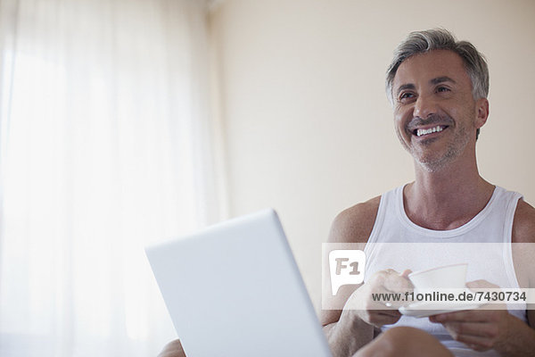 Lächelnder Mann trinkt Kaffee und benutzt Laptop im Schlafzimmer