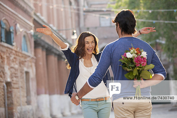 Enthusiastische Frau nähert sich dem Mann mit Blumen hinter dem Rücken.