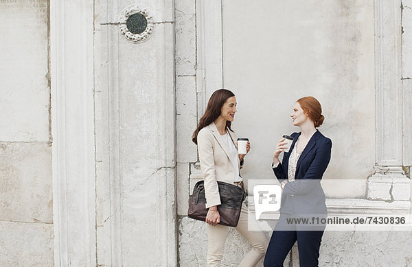 Lächelnde Geschäftsfrauen trinken Kaffee und reden gegen die Gebäudewand