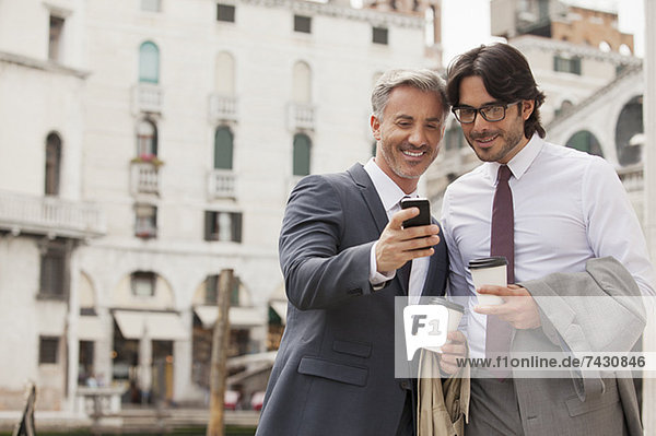 Smiling businessmen with coffee looking down at cell phone in Venice