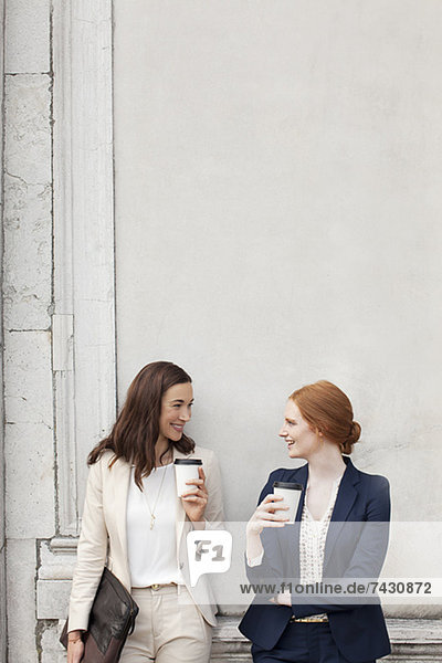 Lächelnde Geschäftsfrauen trinken Kaffee und reden gegen die Gebäudewand