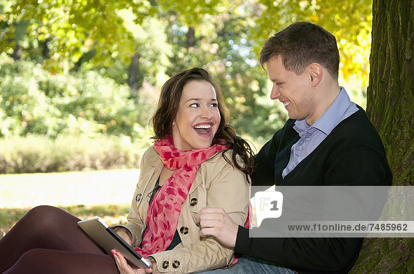 Deutschland  Berlin  Paar sitzend im Herbstpark mit digitalem Tablett  lächelnd