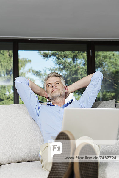 Reifer Mann auf dem Sofa sitzend mit Laptop und Tagträumen