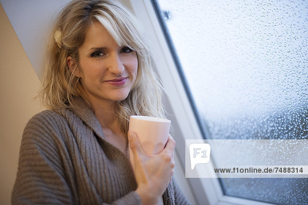 Junge Frau am Fenster stehend mit Kaffee  lächelnd  Portrait
