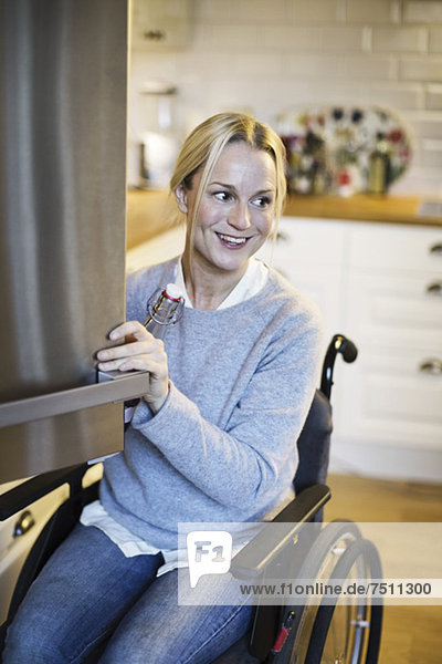 Happy disabled woman with bottle opening refrigerator's door at kitchen