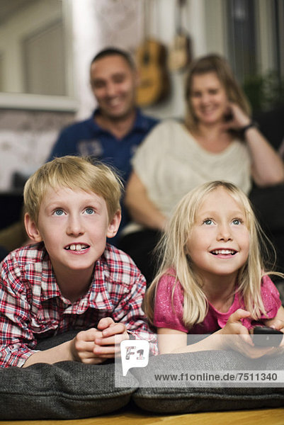 Fröhliche kleine Geschwister beim Fernsehen mit den Eltern im Hintergrund