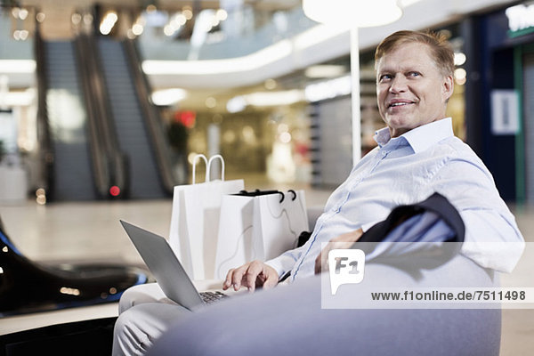 Fröhlicher älterer Mann sitzt auf dem Sofa mit Laptop im Einkaufszentrum