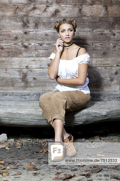 Young woman wearing a white dirndl top and traditional leather pants sitting on a wooden bench  dirndl look