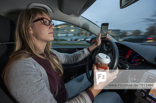 Junge Frau fährt Auto in der Innenstadt  telefoniert mit dem Handy während der Fahrt  hält gleichzeitig einen Kaffeebecher fest