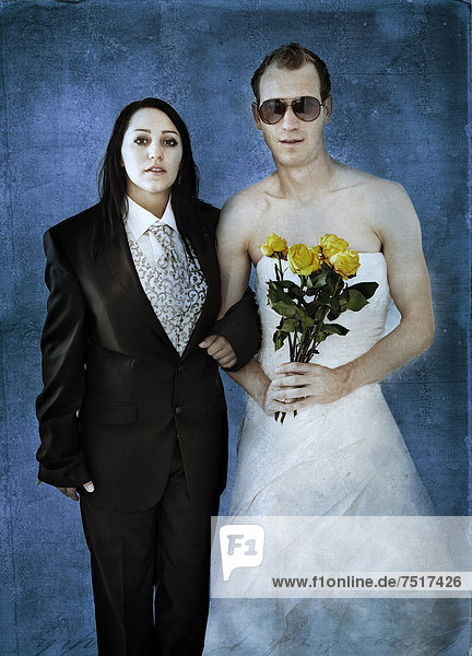 Braut im Hochzeitsanzug und Bräutigam im Hochzeitskleid mit Sonnenbrille und gelben Rosen
