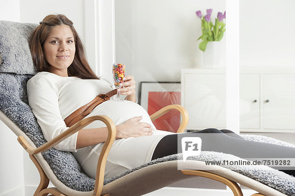 Schwangere liegt auf einem Sessel und hält ein Glas gefüllt mit bunten Süßigkeiten auf ihrem Bauch