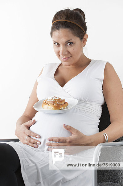 Schwangere Frau hält die Hände auf ihren Bauch und ein Teller mit einem süßen Brötchen balanciert auf ihrem Bauch