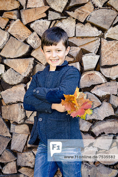 Boy holding autumn leaves  standing in front of stacked firewood