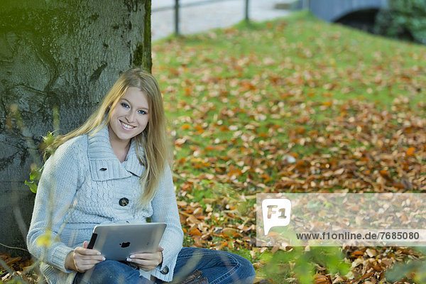 Junge Frau lehnt an einem Baum im Park mit einem Tablet-PC