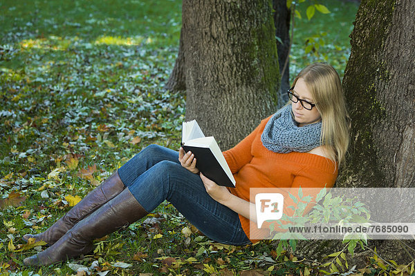 Junge Frau mit Brille sitzt im Park und liest ein Buch