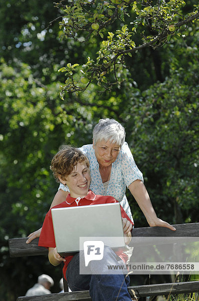 Grandson and grandmother looking together at a laptop computer on a park bench