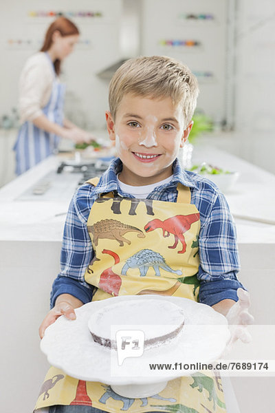 Boy covered in flour holding cake in kitchen