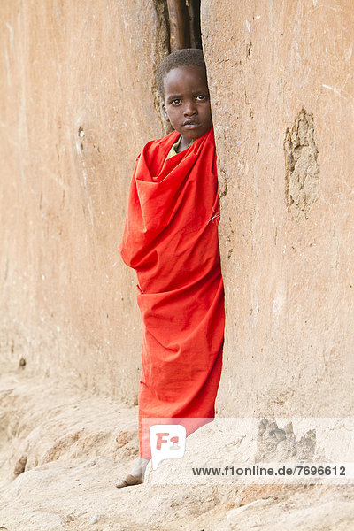 Maasai child wearing traditional dress looking out of a mud hut