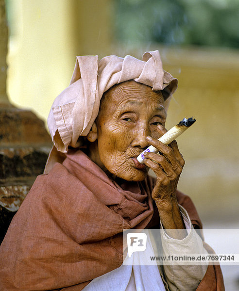 Elderly Burmese woman smoking a Cheerot cigar and wearing a headscarf  beggar  Mingun