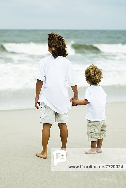 Cute Young Boys On The Beach  Looking At The Ocean Holding Hands.