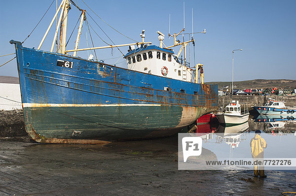 UK  Ireland  County Kerry  Iveragh Peninsula  Fisherman standing on wet shore beside large fishing vessel  Portmagee