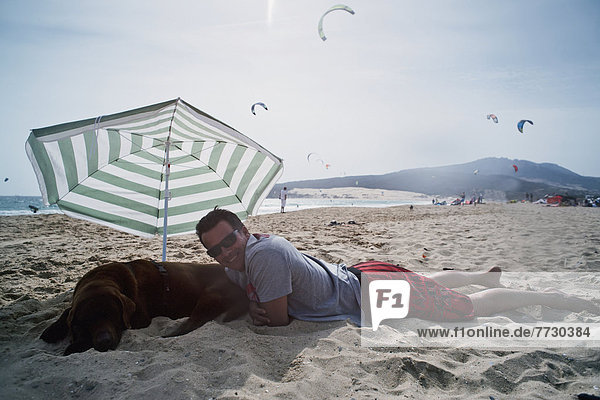 liegend liegen liegt liegendes liegender liegende daliegen Mann Strand Regenschirm Schirm unterhalb Hund Sonnenschirm Schirm