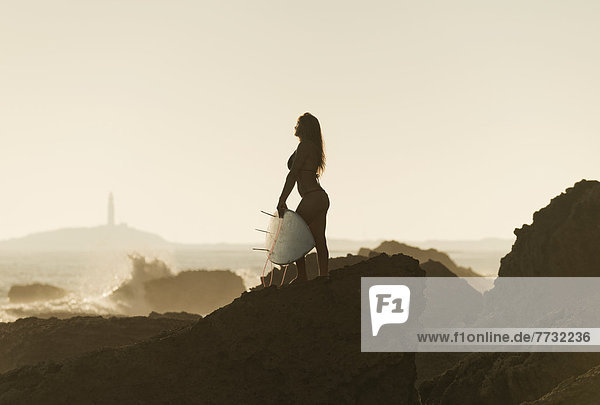 Silhouette of a female surfer holding her surfboard while standing on a rock at the coast  andalusia spain