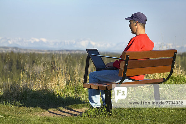 Man Sitting On A Bench In A Park Working On A Laptop With Snow Covered Mountains And Blue Sky In The Background  Calgary  Alberta  Canada