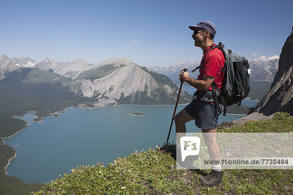 Male Hiker Standing On A Mountain Ridge Overlooking An Emerald Lake And Mountains Below With Blue Sky In Kananaskis Provincial Park  Alberta Canada