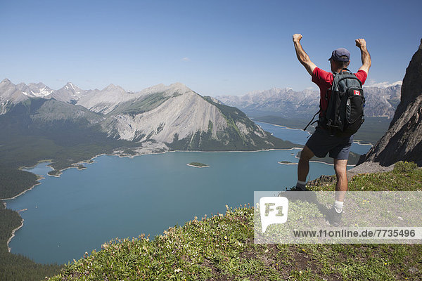 Male Hiker Standing With Arms Up On A Mountain Ridge Overlooking An Emerald Lake And Mountains Below With Blue Sky In Kananaskis Provincial Park  Alberta Canada
