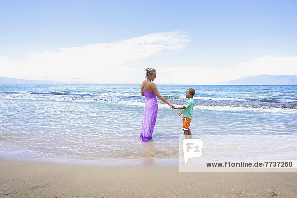 A mother and son hold hands while standing in the shallow water at the ocean Hawaii united states of america