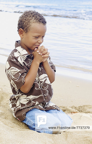 A young boy with hands folded in prayer by the ocean Hawaii united states of america