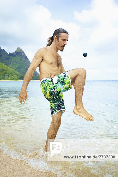 A man bumps a hackey sack with his knee while standing in the shallow water of the ocean Hawaii united states of america