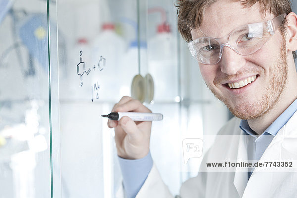 Deutschland  Porträt eines jungen Wissenschaftlers  der chemische Gleichungen auf Glas schreibt  lächelnd