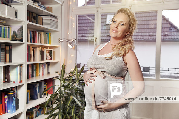 Schwangere Frau steht im Wohnzimmer und hält ihren Bauch  lächelnd  Portrait