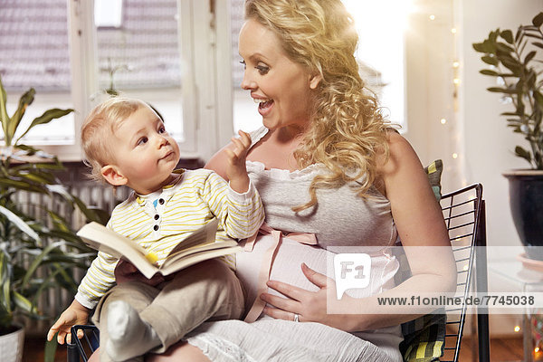 Germany  Bonn  Pregnant mother reading book to son in living room  smiling