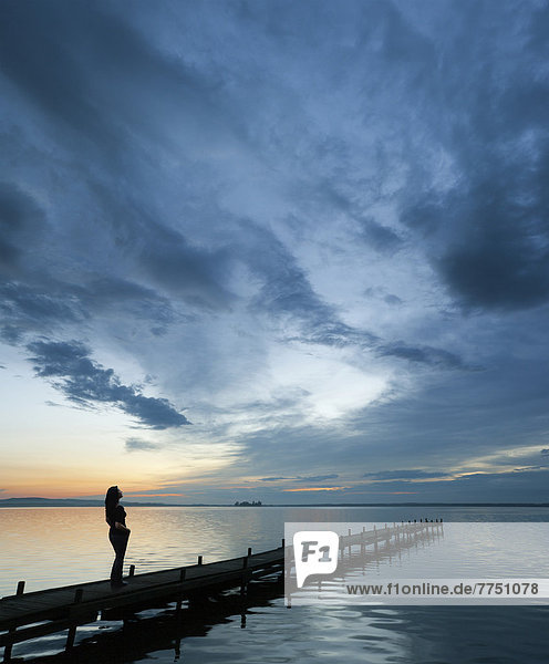 Frau steht auf einem Holzsteg am Seeufer den reizvollen Abendhimmel bewundert