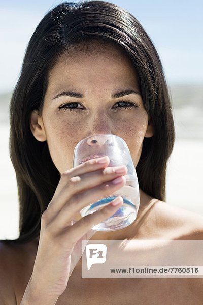 Braunhaarige junge Frau am Strand  die ein Glas Wasser trinkt.