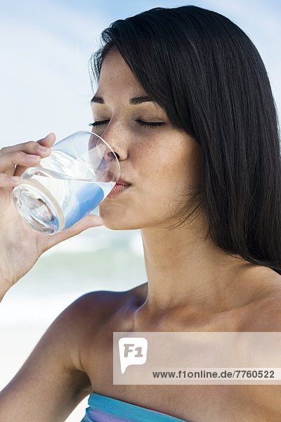 Braunhaarige junge Frau am Strand  die ein Glas Wasser trinkt.