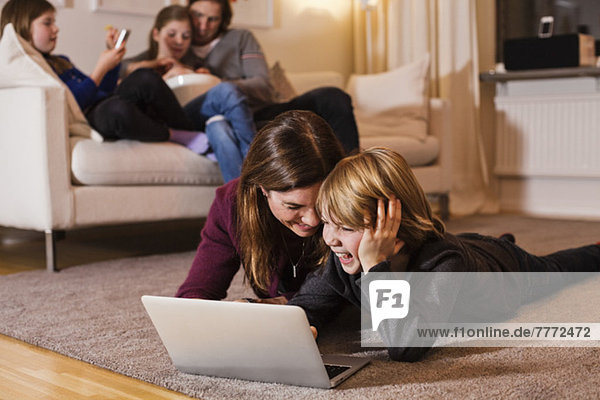 Glückliche Mutter und Sohn mit Laptop auf dem Boden mit Familie auf Sofa im Wohnzimmer sitzen
