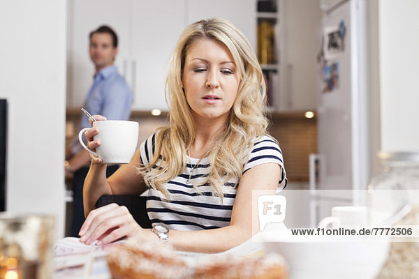 Junge Frau hält Kaffeetasse und liest Zeitung mit Mann im Hintergrund