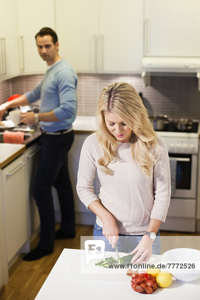 Frau schneidet Gemüse an der Küchentheke mit Mann im Hintergrund